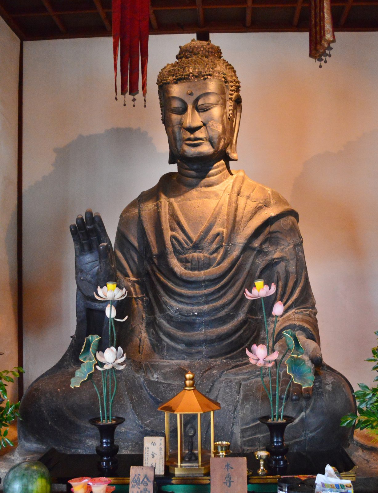 一座寺庙佛堂里，一尊高大的铜坐佛，右手举起掌心向外，面部有明显的修补痕迹，身体颜色斑驳；佛前供着莲花、灯和小塔。