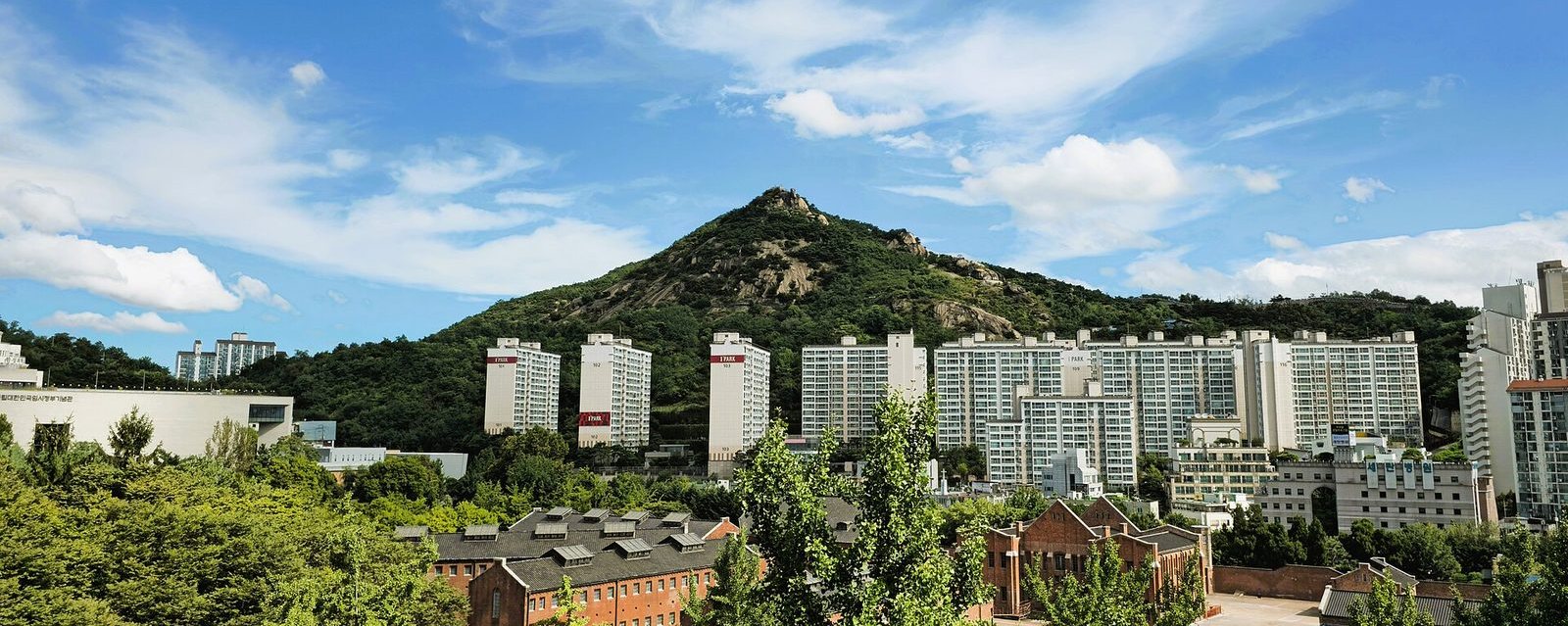 今天的仁王山，从首尔西大门一带望去：一座不算高的山，山顶露出灰白的花岗岩，山坡长满绿树；山脚下是一排排高层公寓和红砖建筑。